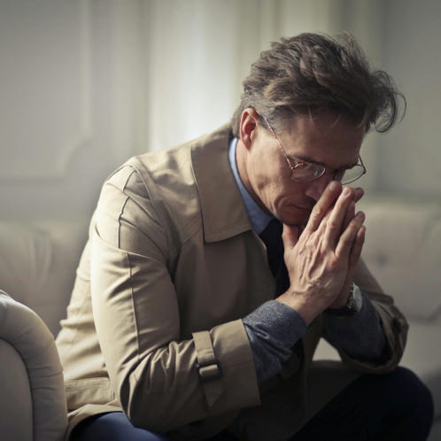 Side view of upset male entrepreneur in formal wear sitting on couch in modern living room and contemplating about financial problems while leaning on hands and looking down