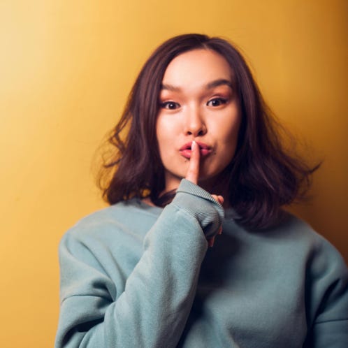 Young Asian female in warm clothing doing shh gesture with finger on lips and looking at camera while standing near orange wall