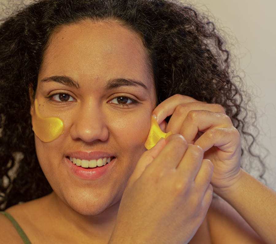 Optimistic ethnic female doing skin care procedure with gel eye patches and looking at camera with toothy smile