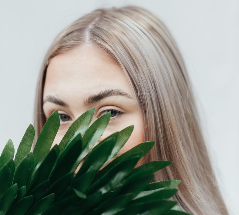 Young female covering half face with green branch of fresh exotic plant against white background